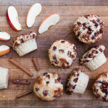 Vegan apple cinnamon muffins spread out on a wooden cutting board with sliced apples and cinnamon sticks.