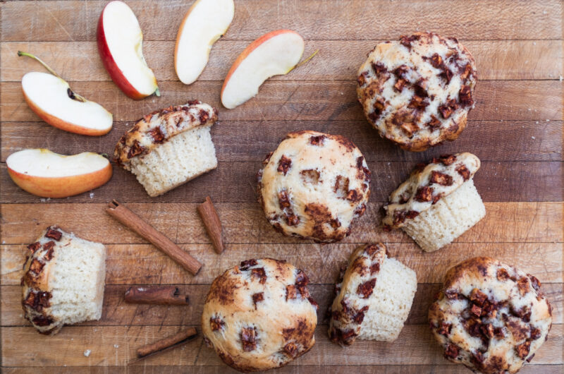 Vegan apple cinnamon muffins spread out on a wooden cutting board with sliced apples and cinnamon sticks.