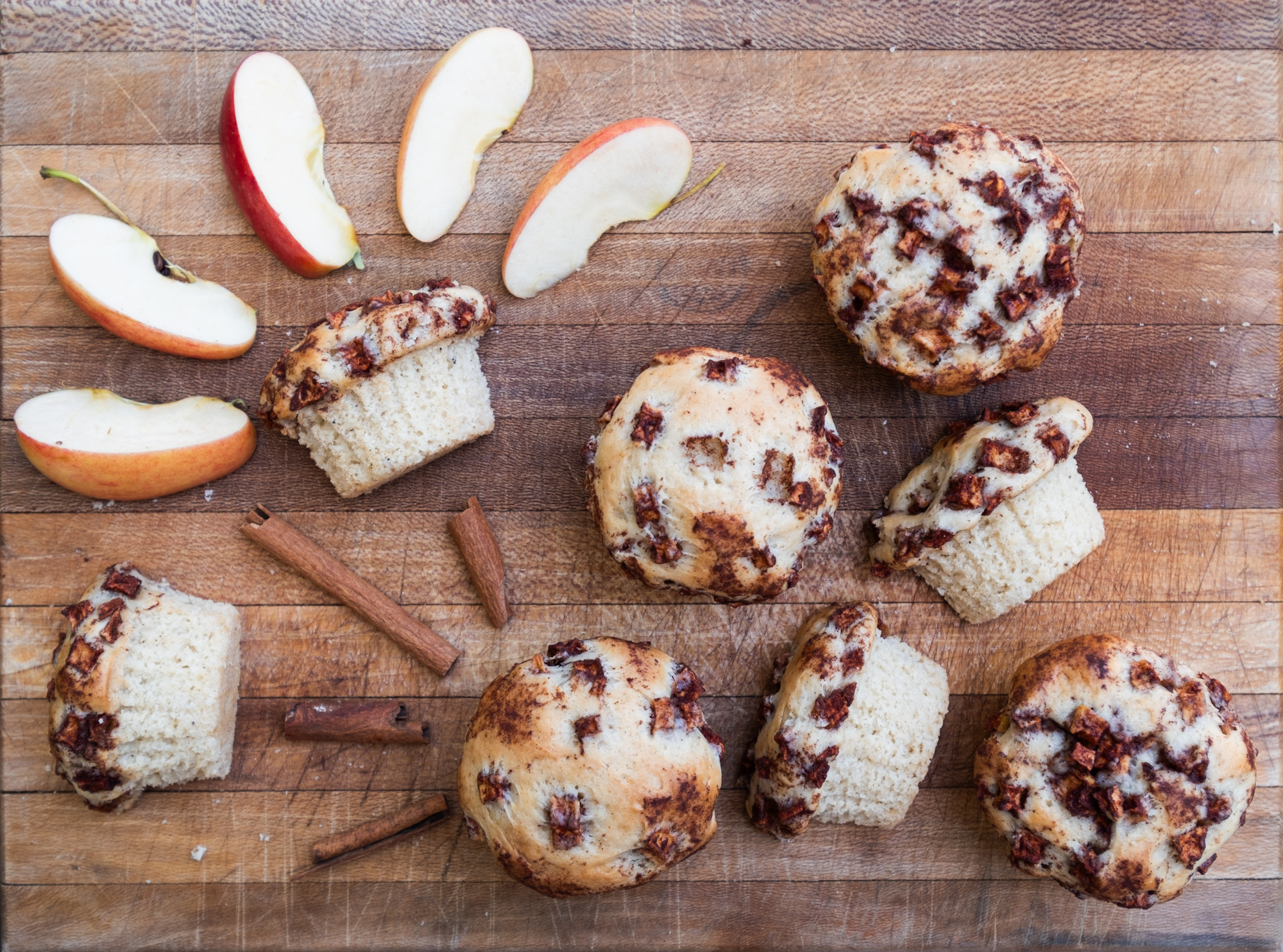 Vegan apple cinnamon muffins spread out on a wooden cutting board with sliced apples and cinnamon sticks.